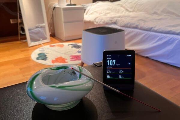 an air purifier in a bedroom next to an air quality monitor showing high levels of indoor air pollution