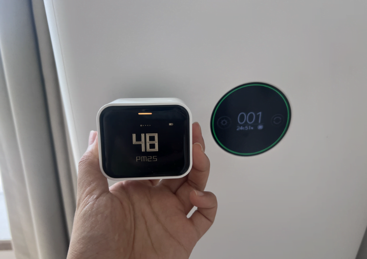 a human hand holding an air quality monitor next to a built in sensor of an air purifier